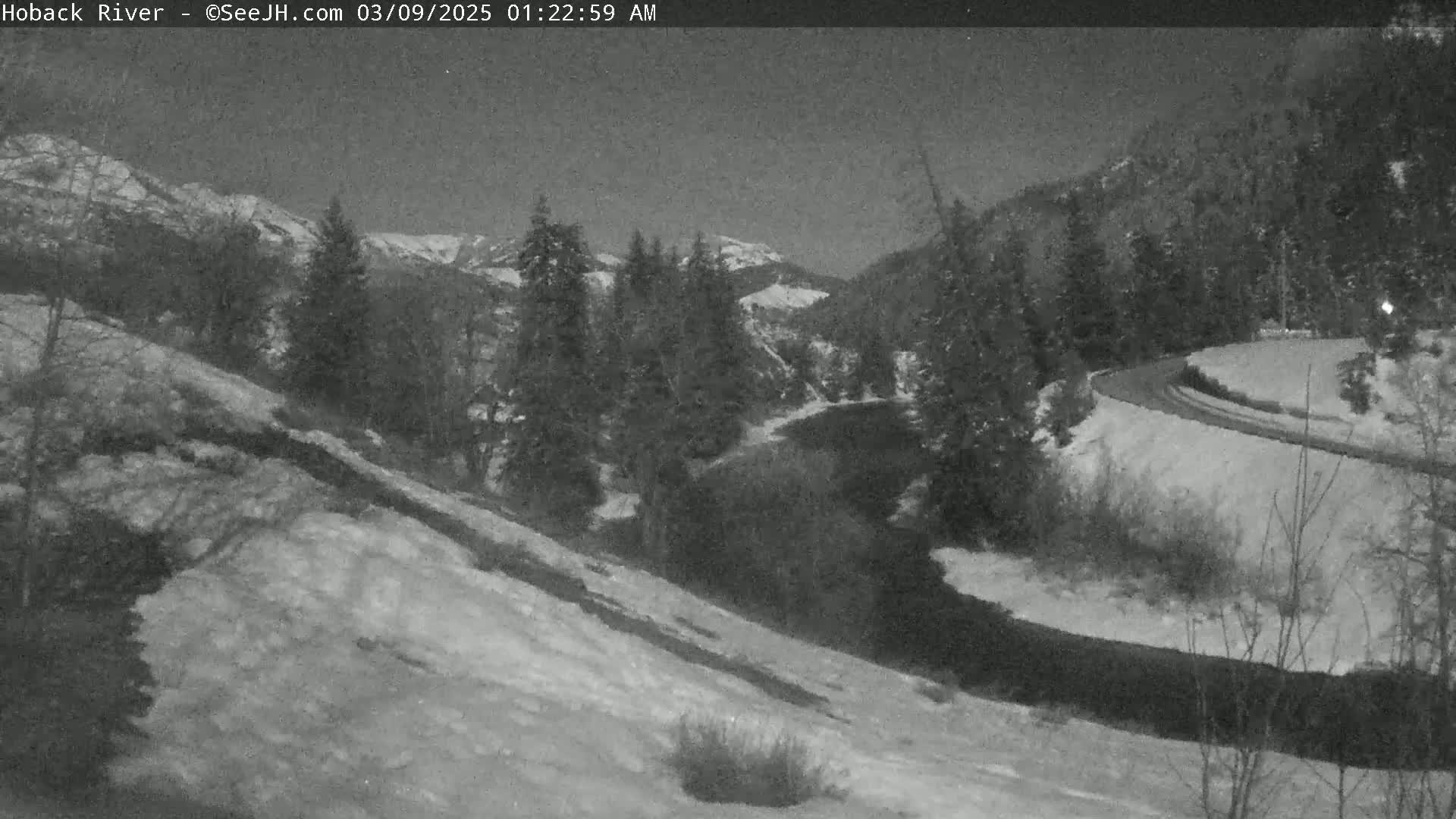 A nighttime view of a snow-covered landscape with a dark river winding through a valley, bordered by dark evergreen trees and snow-covered hills under a clear sky.