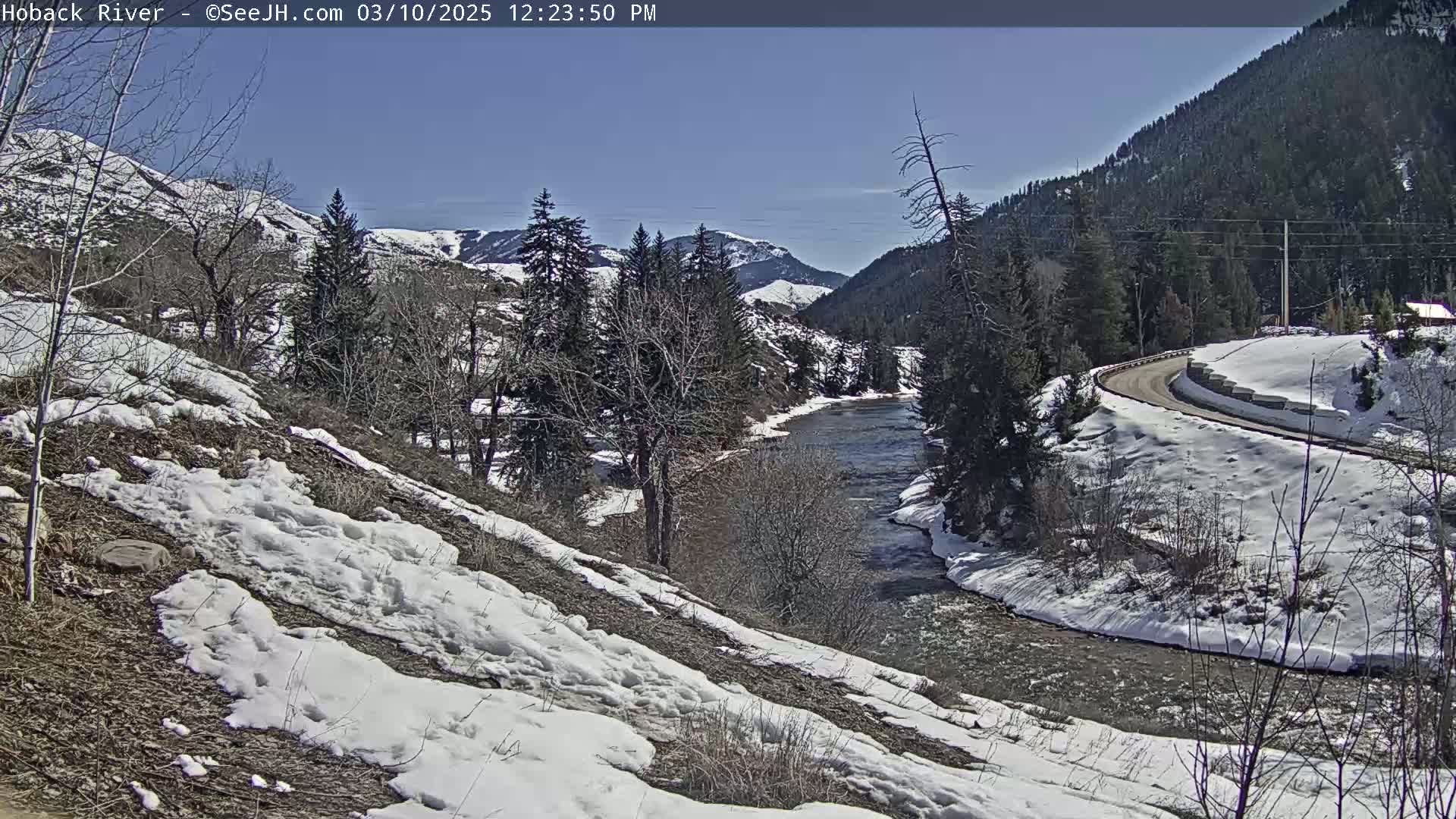 A snow-dusted river winds through a valley under a clear blue sky, flanked by snow-covered hillsides and evergreen trees.
