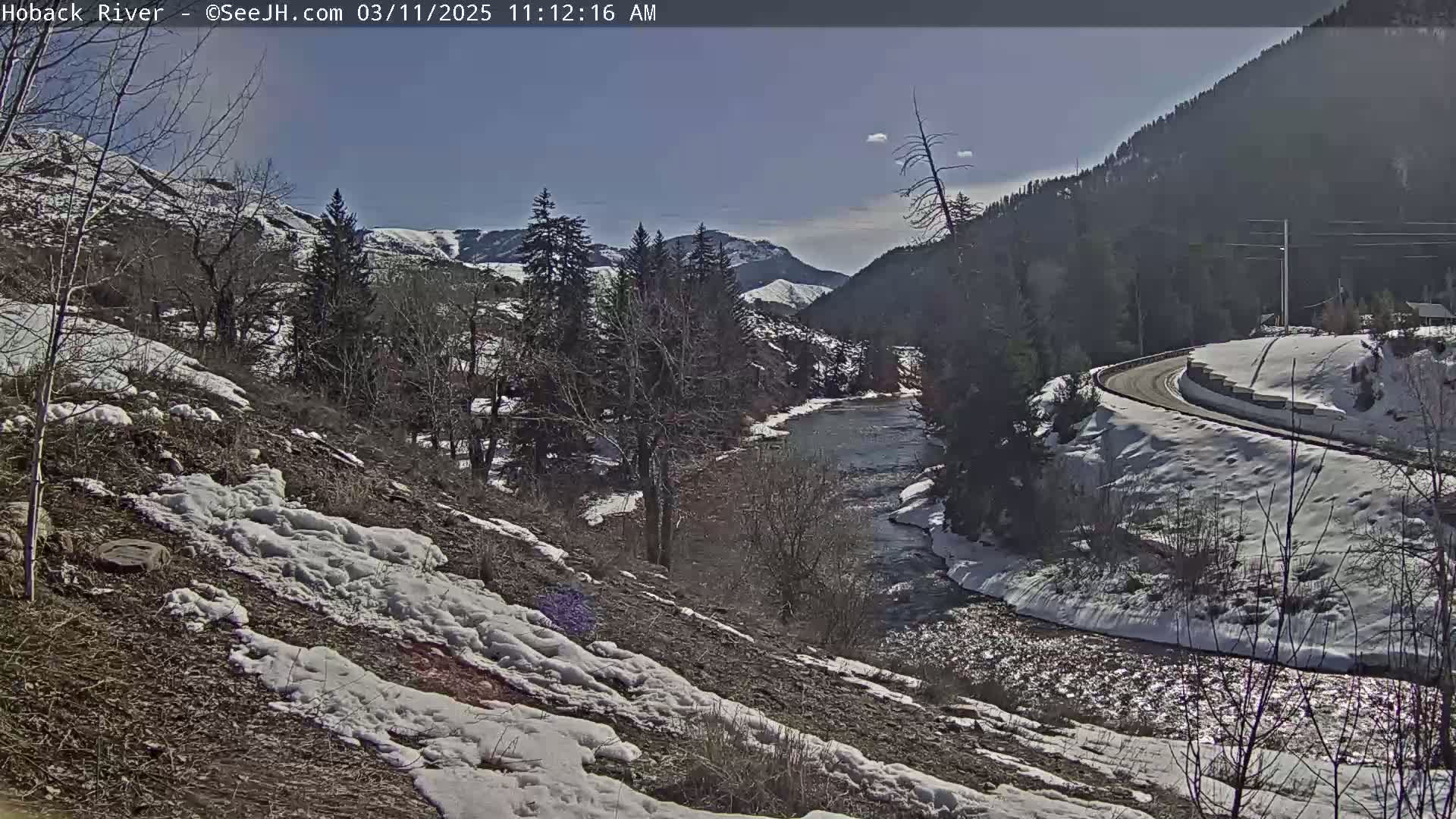 A partly sunny day shows a winding river flowing through a snow-dusted valley, with a road curving along one side.