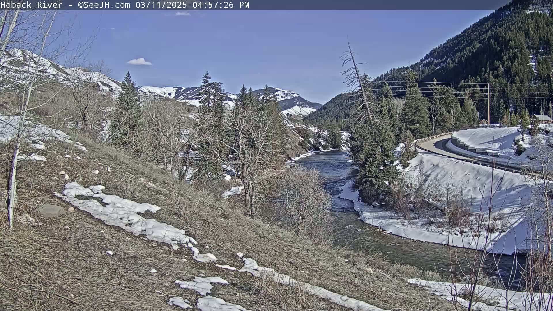 A river winds through a snow-dusted valley under a mostly sunny sky, with a road curving along the opposite bank, surrounded by leafless trees and snow-covered hills.