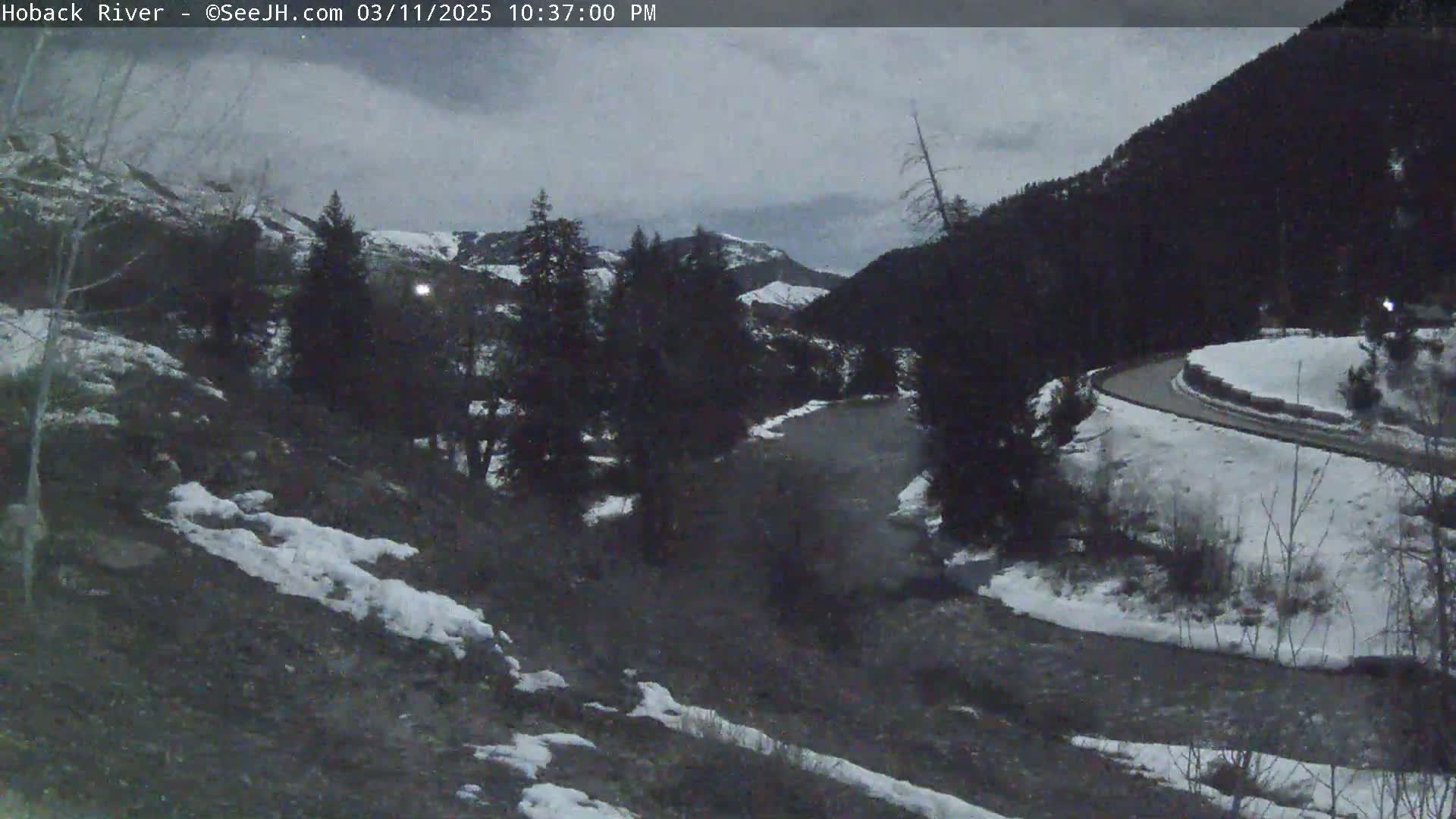 A dark, overcast evening scene shows a river winding through a snow-covered valley with dark evergreen trees and a road curving along the hillside.