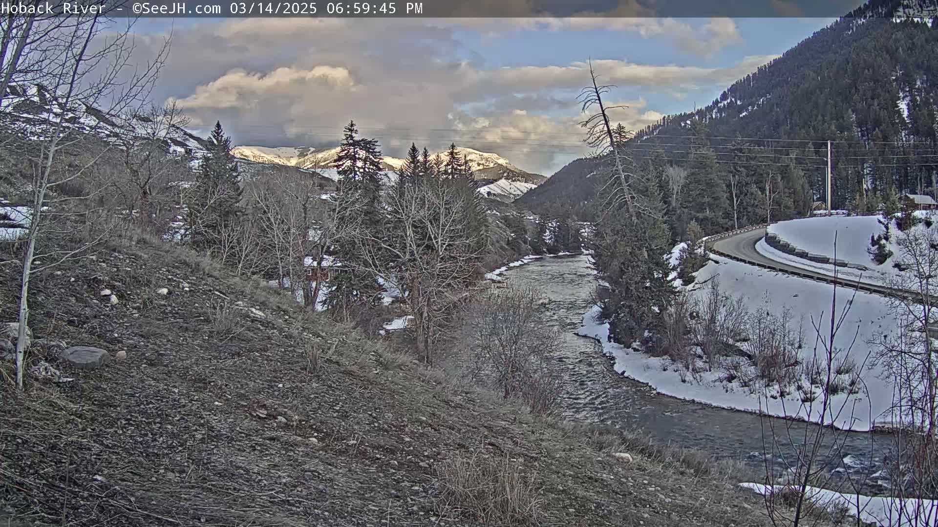 A river flows through a snow-covered valley under a partly cloudy sky, with a road winding along the riverbank and mountains in the background.
