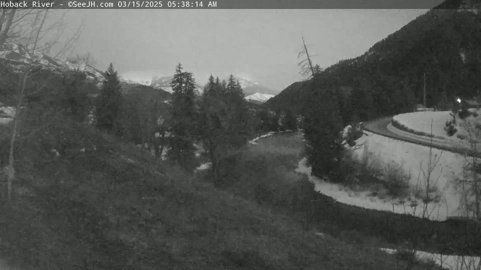 A winding road curves through a snow-covered landscape, alongside a river, under a gray, overcast sky, with evergreen trees and mountains in the background.