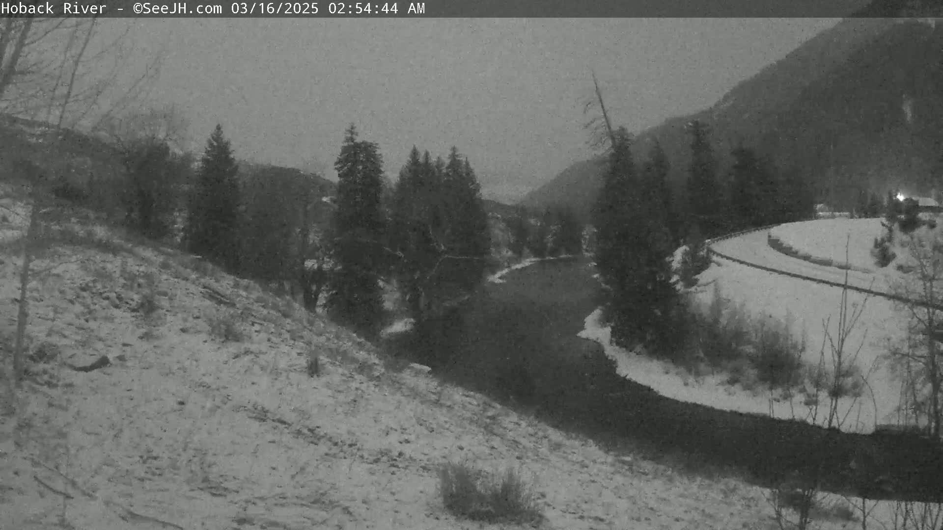 A snow-covered landscape shows a dark river winding through a valley of evergreen trees under a gray, overcast sky.