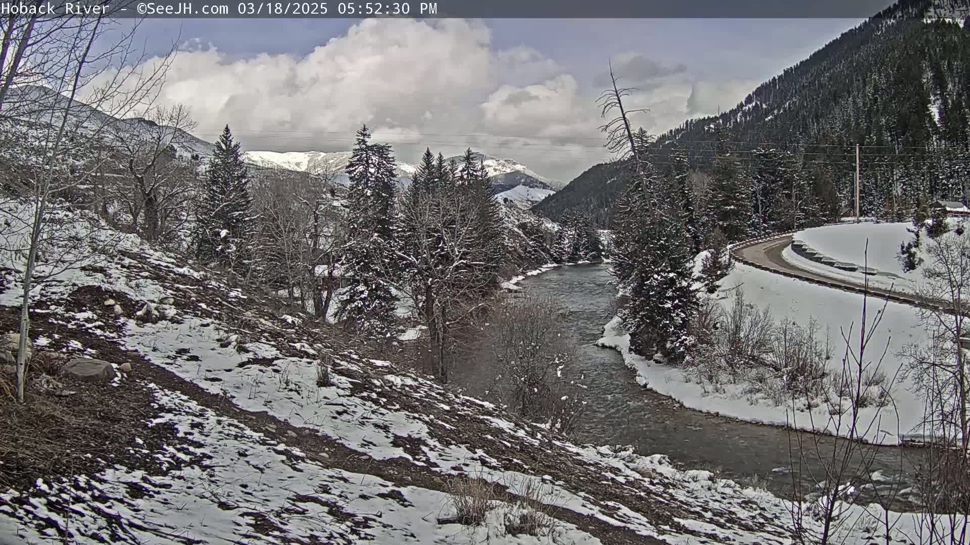 A snow-covered river flows through a valley of snow-dusted evergreen trees and mountains under a partly cloudy sky.