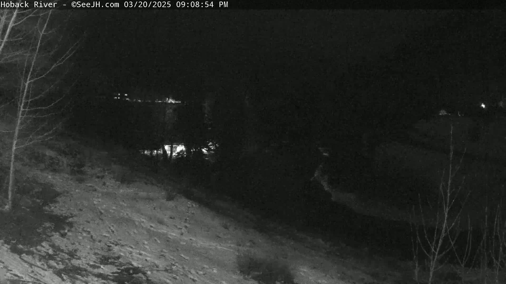 A snow-covered hillside slopes down toward a dark, nighttime riverbank under a clear, dark sky.