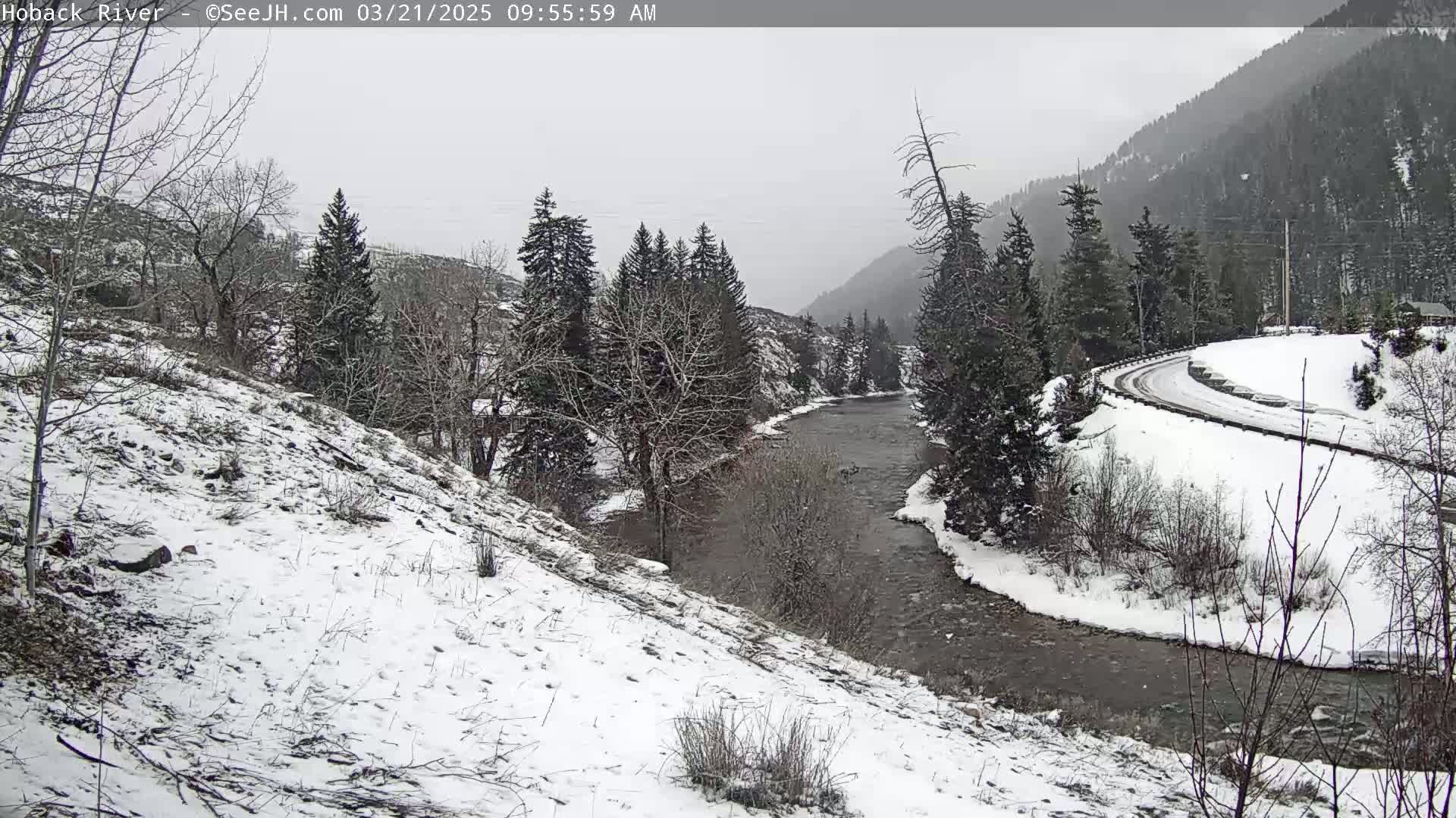A snow-covered landscape reveals a river winding through a valley, with a road curving along a snow-covered hillside in the background, under an overcast sky.