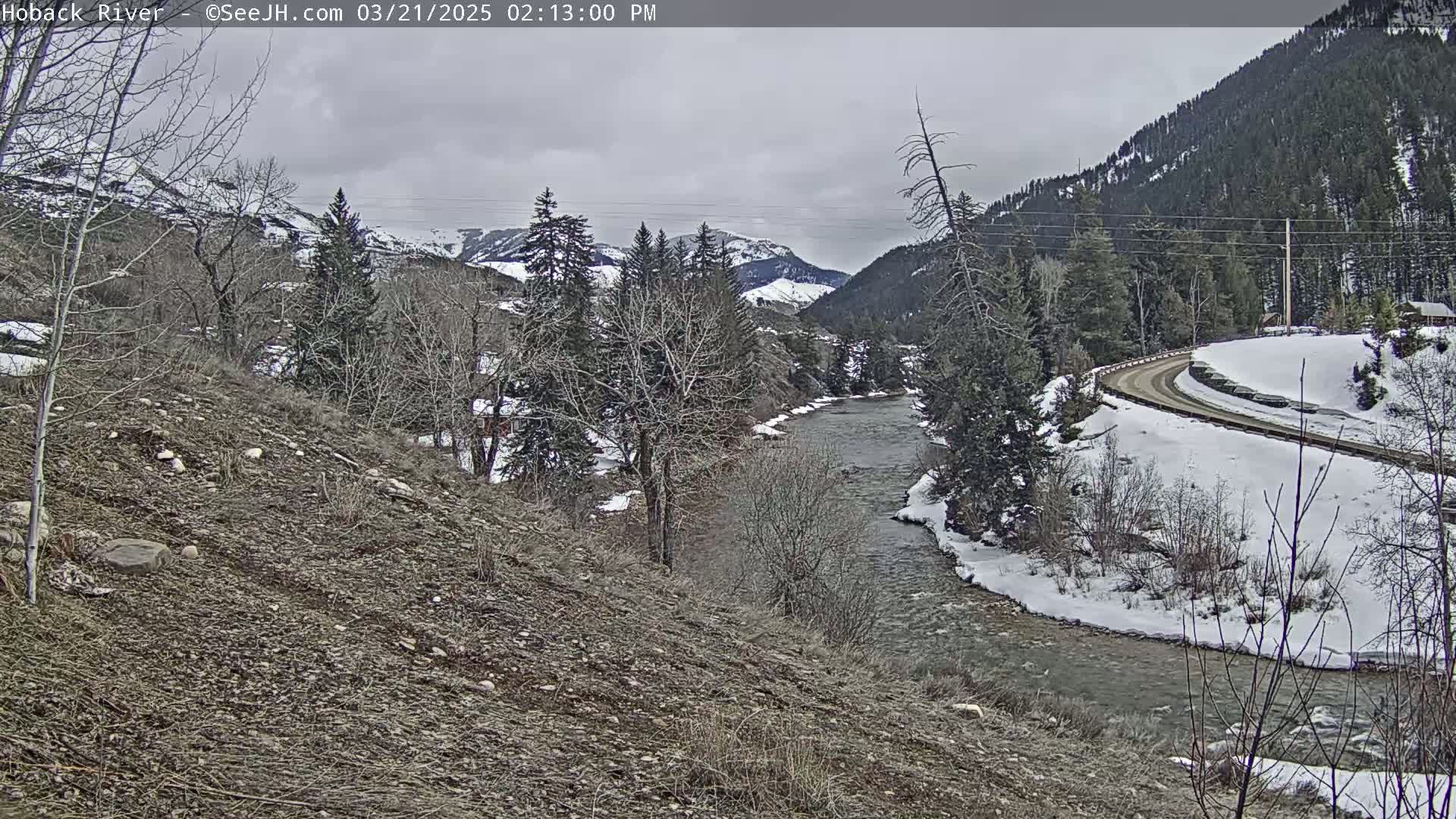 A river flows through a snow-covered valley under an overcast sky, with bare trees and a winding road visible along the banks.