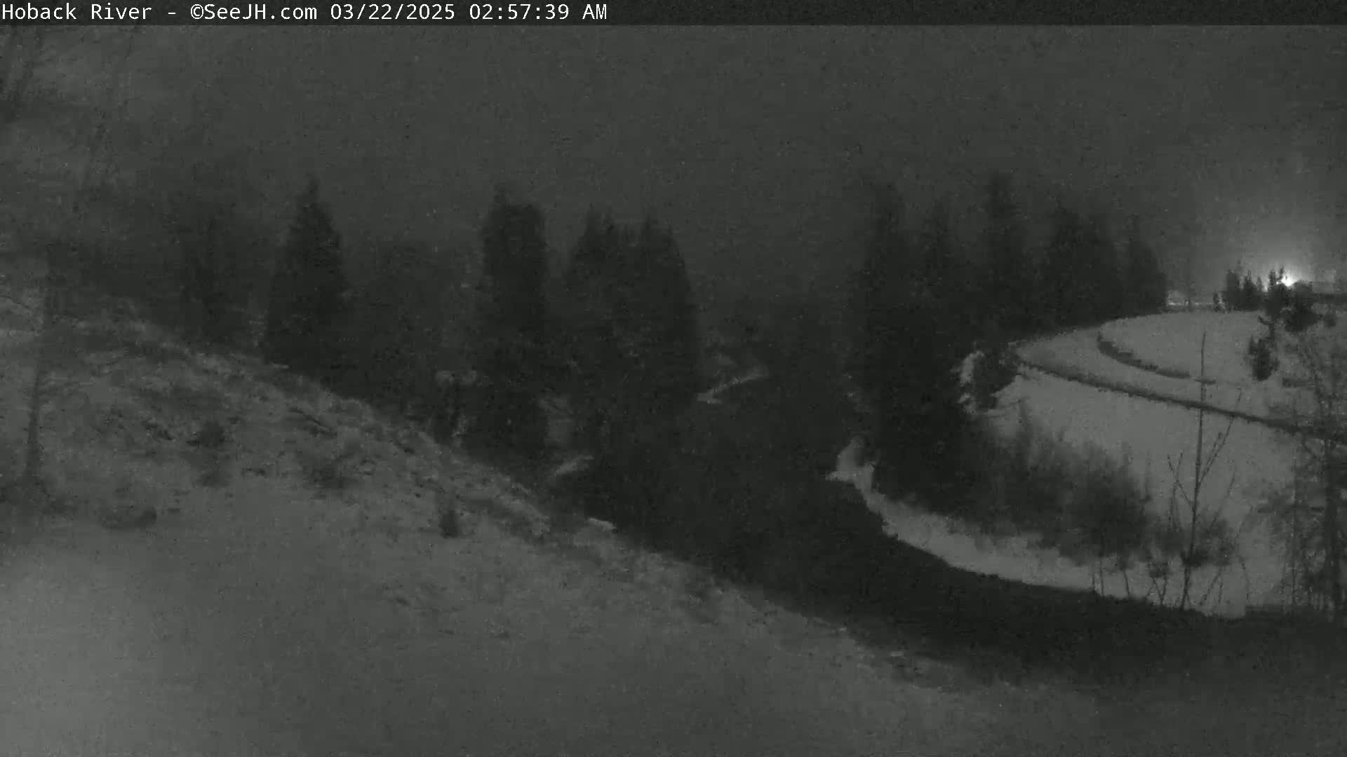 A dark, snowy nighttime scene shows a river winding through a snow-covered landscape with a line of dark trees in the background.