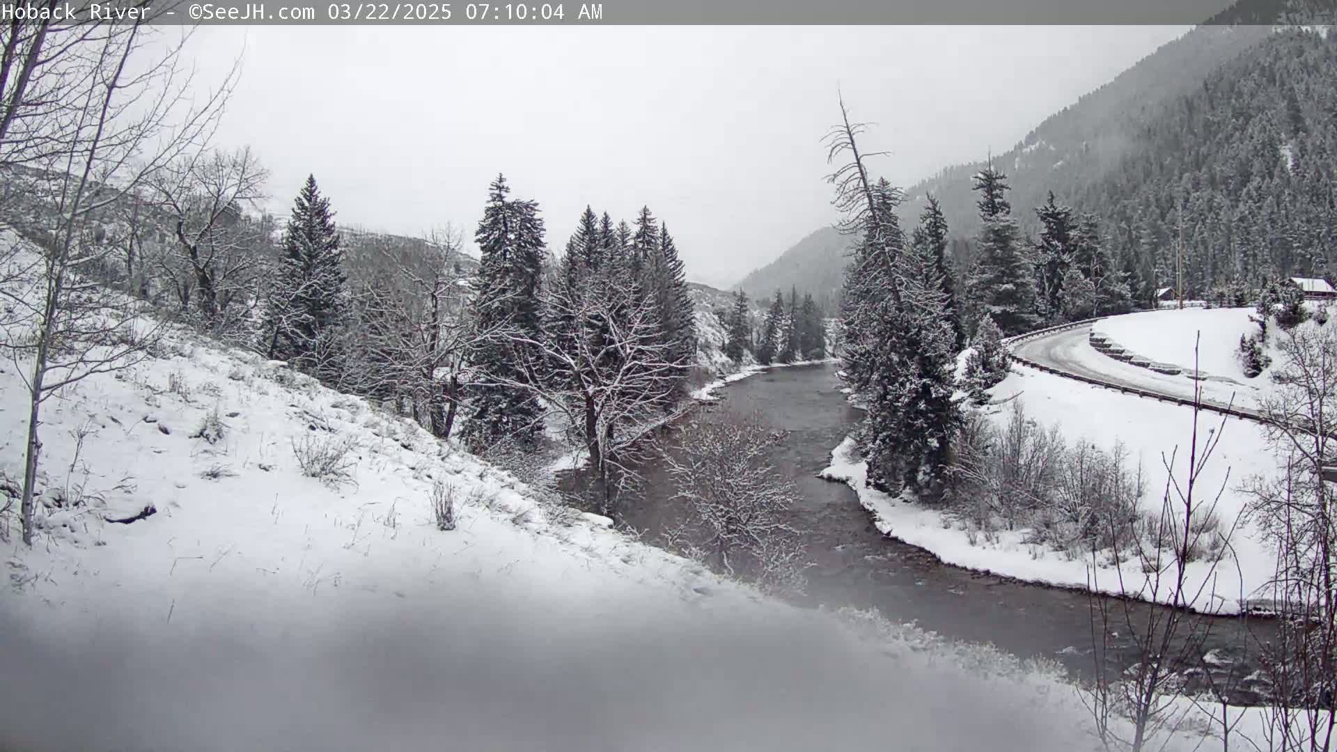 A snow-covered landscape shows a river winding through a valley with snow-laden evergreens and a road curving along a hillside in the background, under an overcast sky.