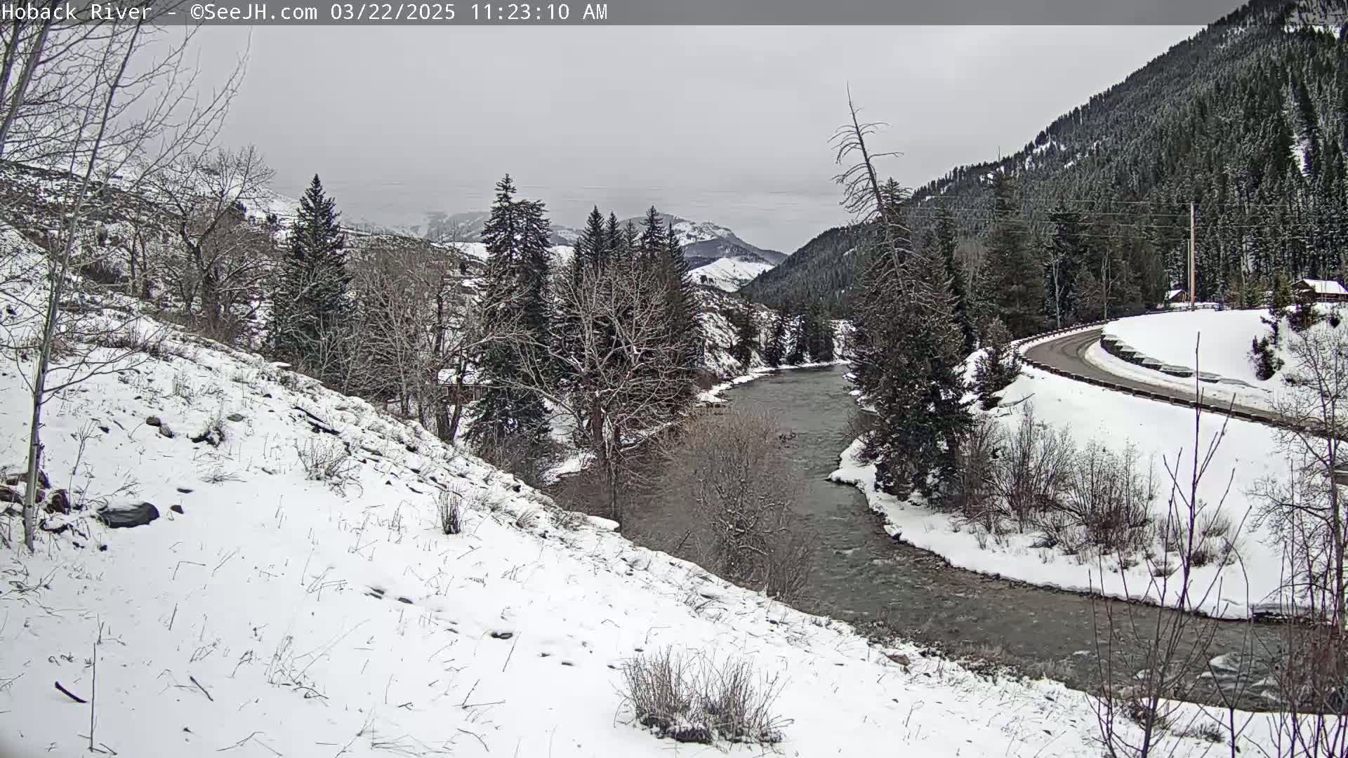 A snow-covered landscape shows a river winding through a valley under an overcast sky, with a road curving along a hillside in the background.