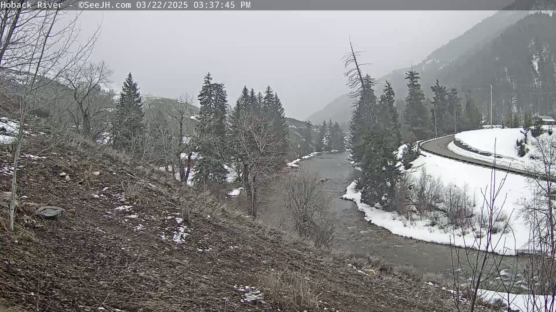 A snow-covered, winding road overlooks a river flowing through a valley of snow-dusted evergreen trees under an overcast sky.