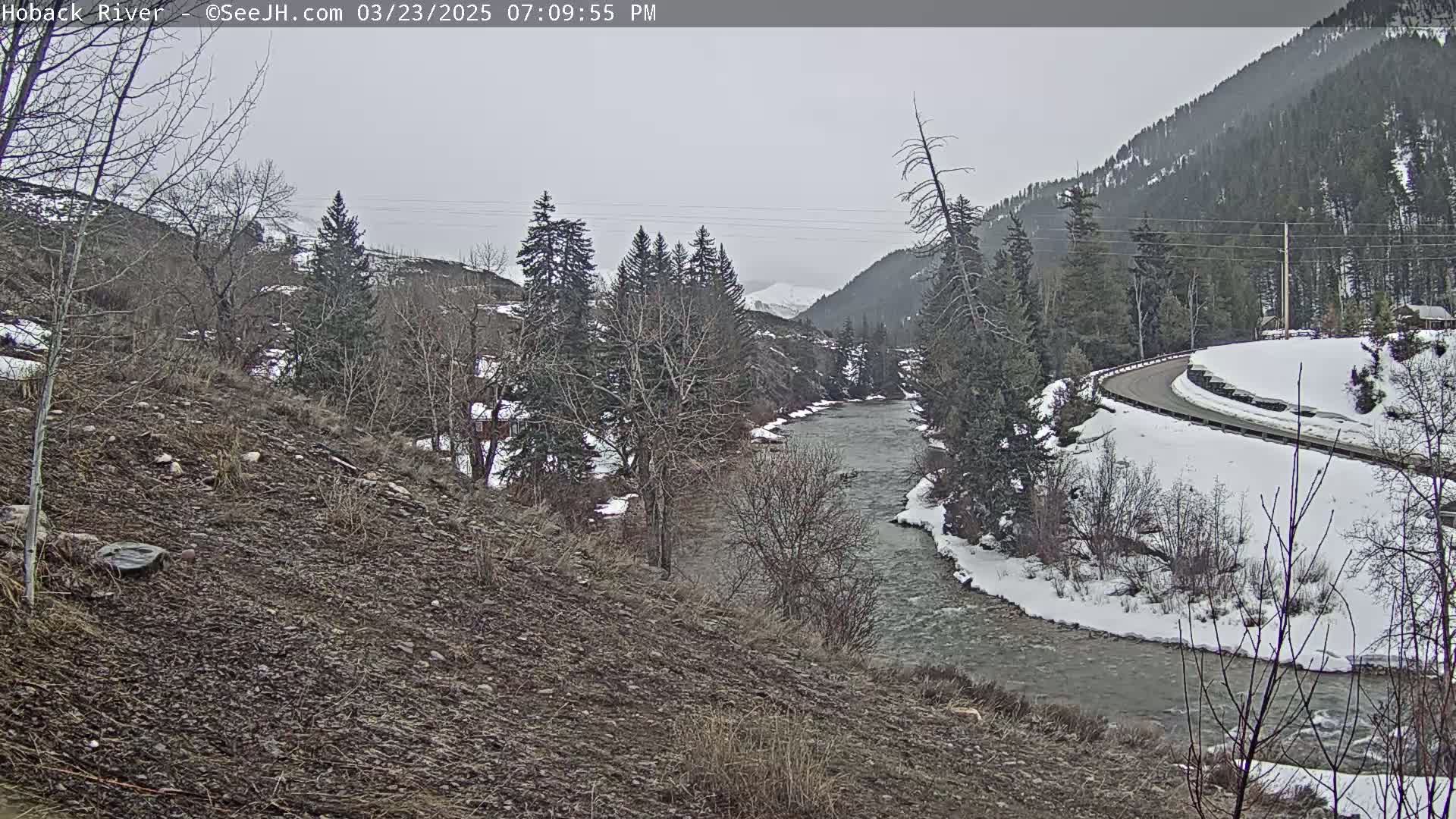 A river flows through a snowy landscape of trees and hills under an overcast sky.