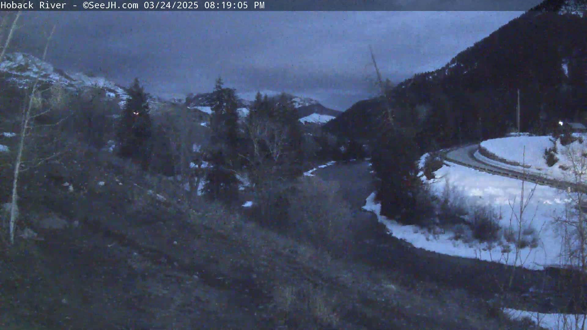 A dark, overcast evening scene shows a river winding through a snow-covered valley between dark mountains.