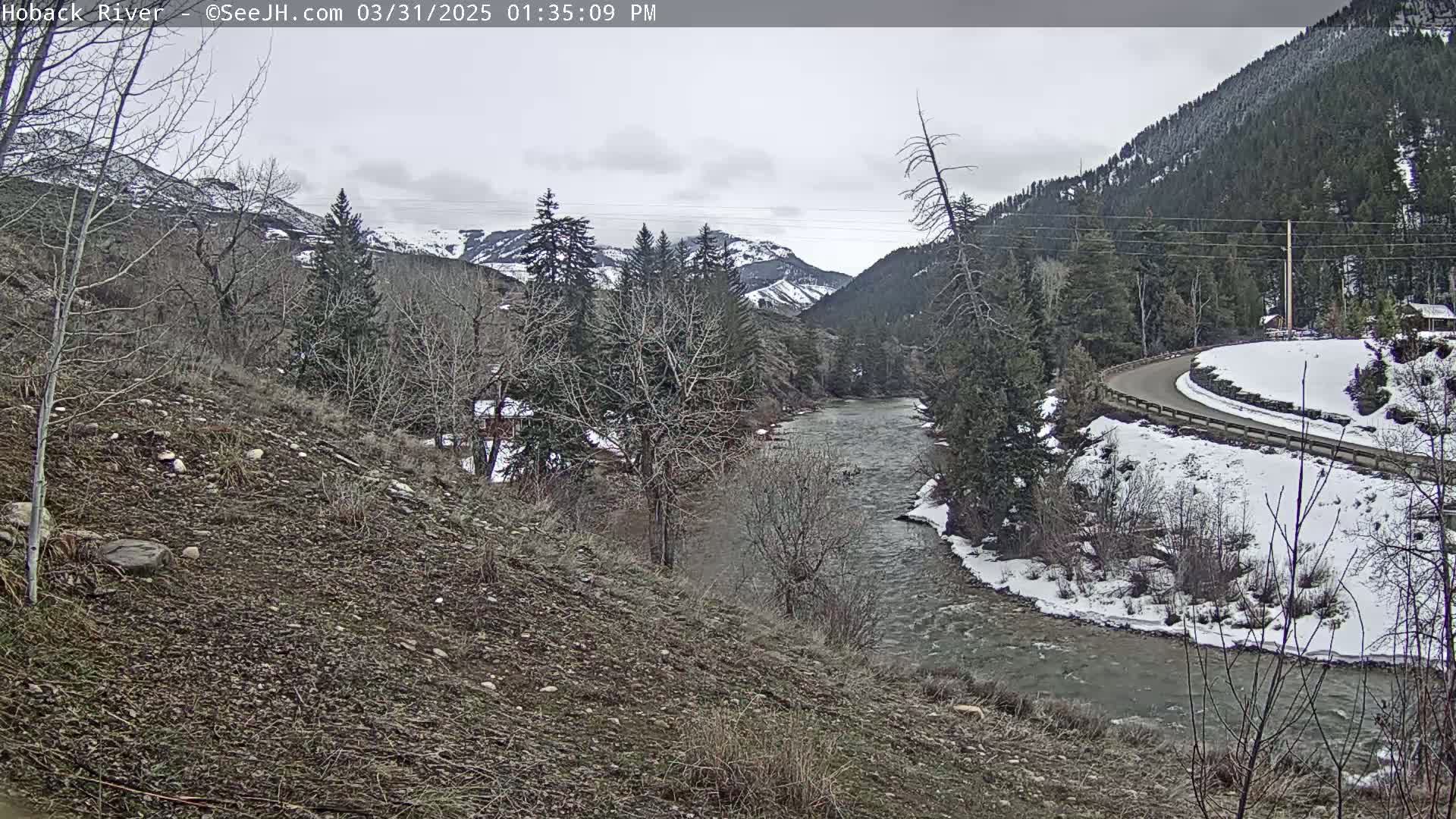 A river flows through a snow-dusted valley under a cloudy sky, with a road curving alongside it.