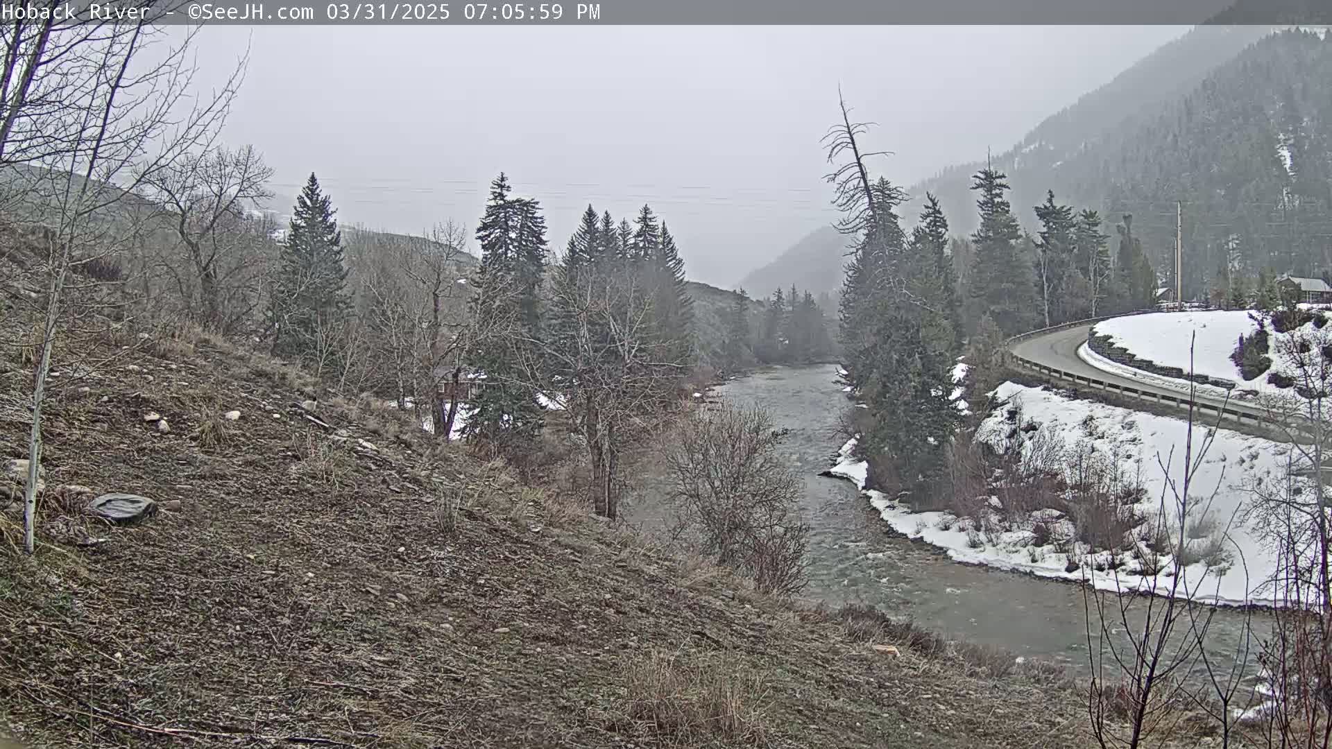 A snow-dusted river flows through a mountainous region, partially covered with snow, under an overcast sky.
