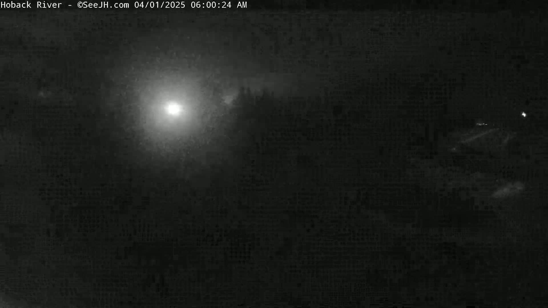 A nighttime, hazy or foggy scene shows a bright light source in the center, possibly a streetlight or moon, with dark, indistinct shapes in the foreground and background.