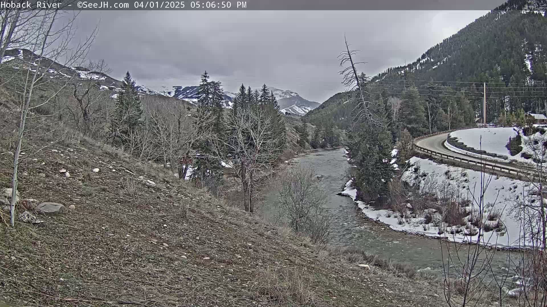 A river flows through a snow-dusted valley under a cloudy sky, alongside a road winding through sparsely wooded hills.