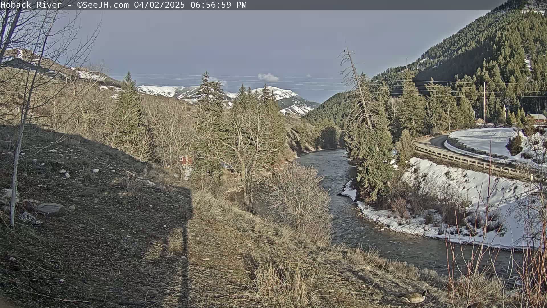 A river flows through a snow-dusted valley, with leafless trees and distant mountains under a mostly clear sky.