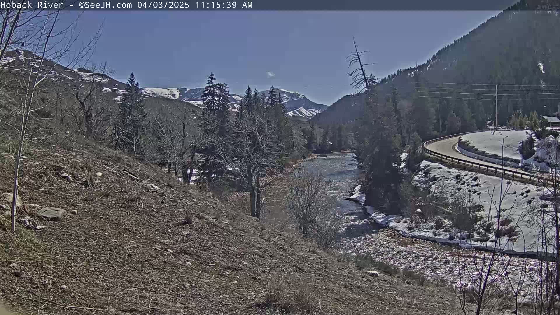 A river flows through a snow-dusted valley under a clear blue sky, with a road curving alongside it.