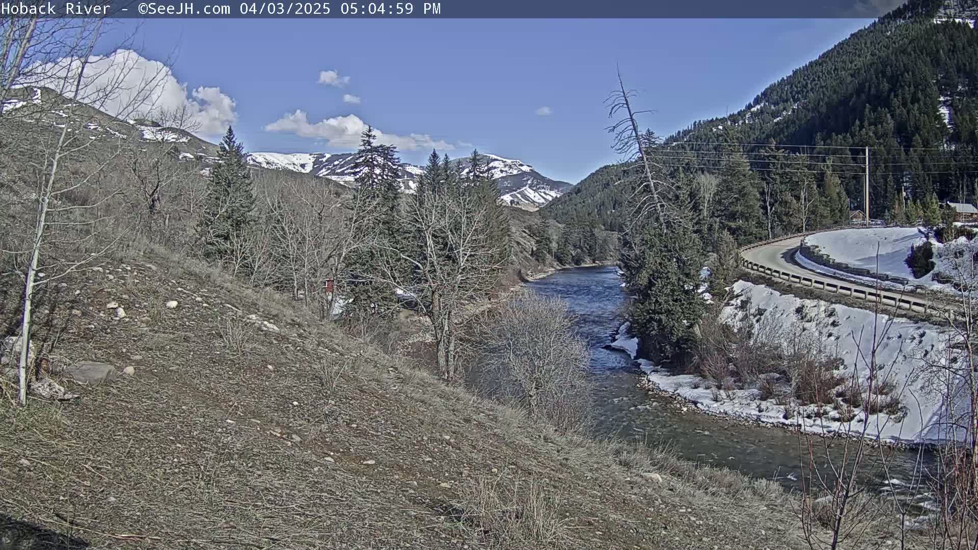 A partly sunny day shows a river flowing through a valley, with snow on the ground and mountains in the background.