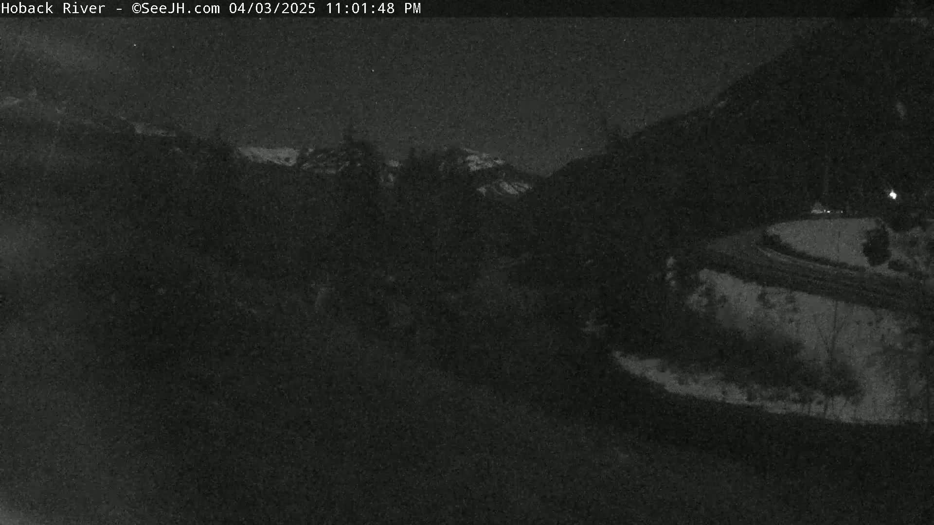 A nighttime, low-light view of a dark, snow-covered road winding through a mountainous landscape under a clear sky.