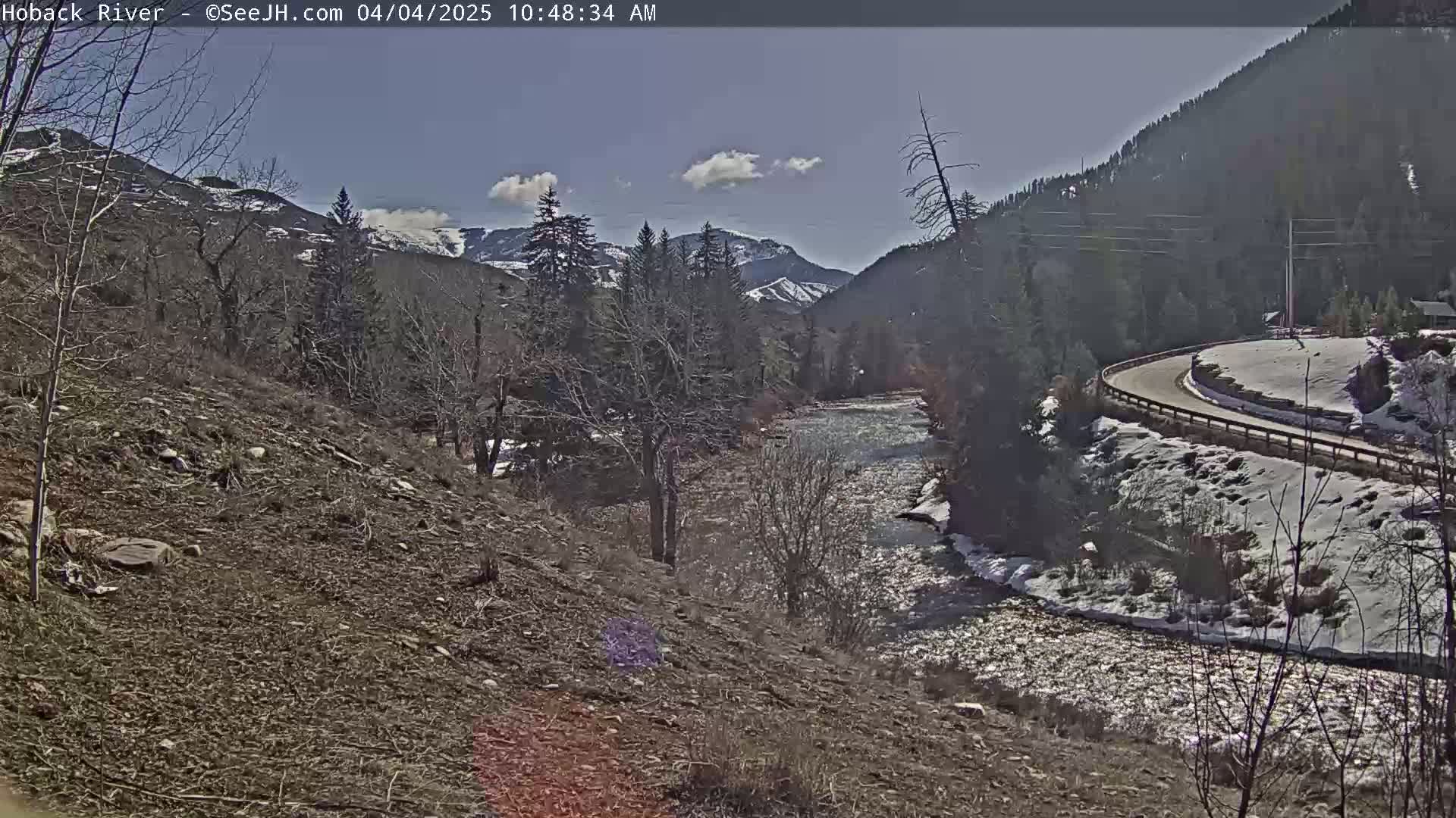 A river flows through a mountainous valley under a mostly sunny sky, with patches of snow remaining on the ground and in the higher elevations.