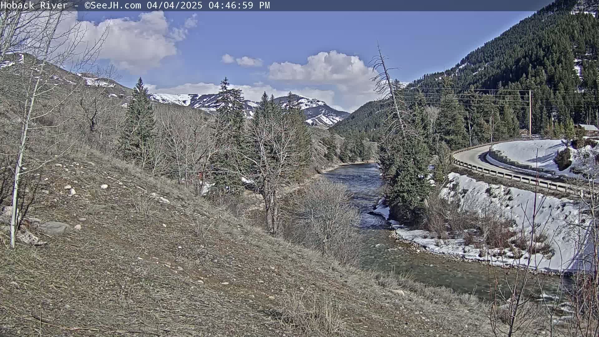 A partly cloudy day shows a river flowing through a valley with snow-capped mountains in the background and some snow along the riverbanks and road.