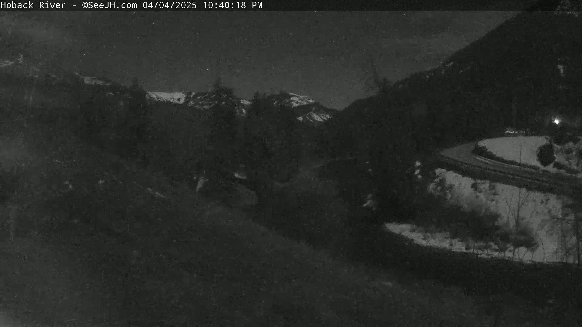 A nighttime view of a snow-covered landscape with a winding road curving through dark, wooded hills under a clear sky.