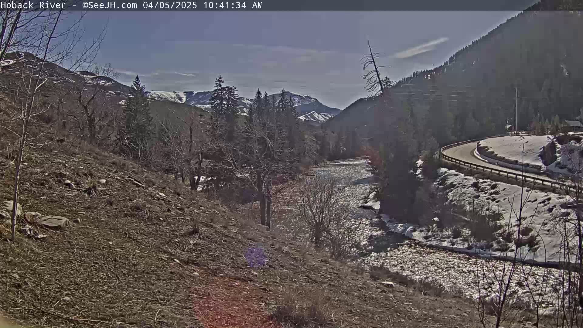 A river flows through a snow-dusted valley under a mostly clear sky, with mountains in the background and a road curving along the opposite bank.