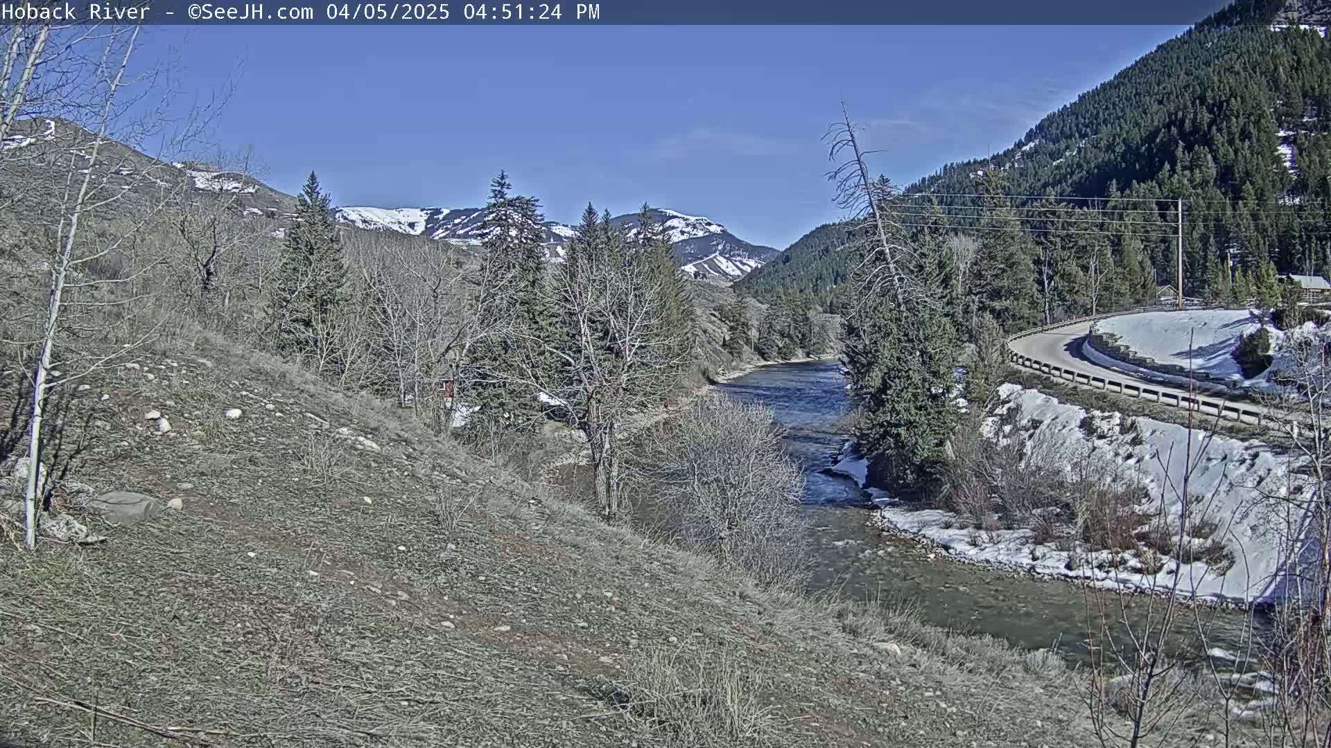 A river flows through a valley on a sunny day, with snow on the ground and mountains in the background.