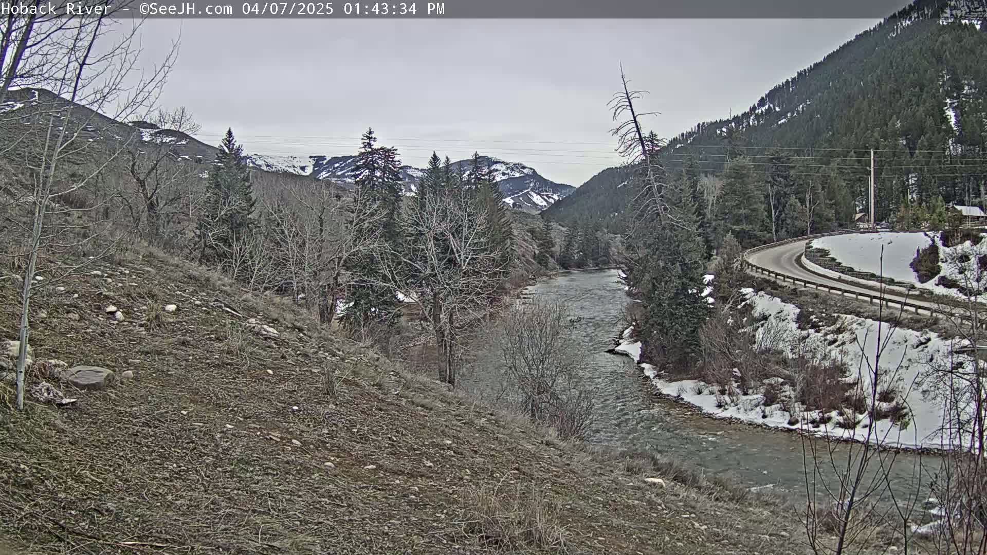 A river flows through a valley under an overcast sky, with snow on the ground and bare trees lining the banks.