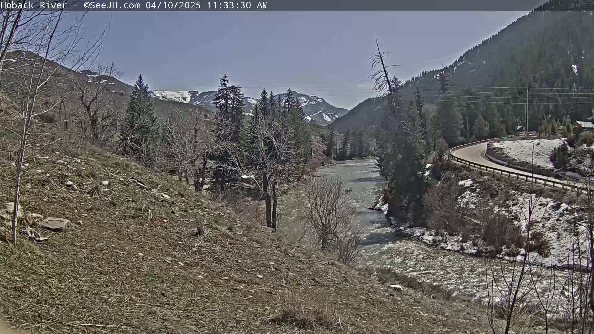 A fast-moving river flows through a mountainous valley on a sunny day with patches of snow on the ground and hillsides.