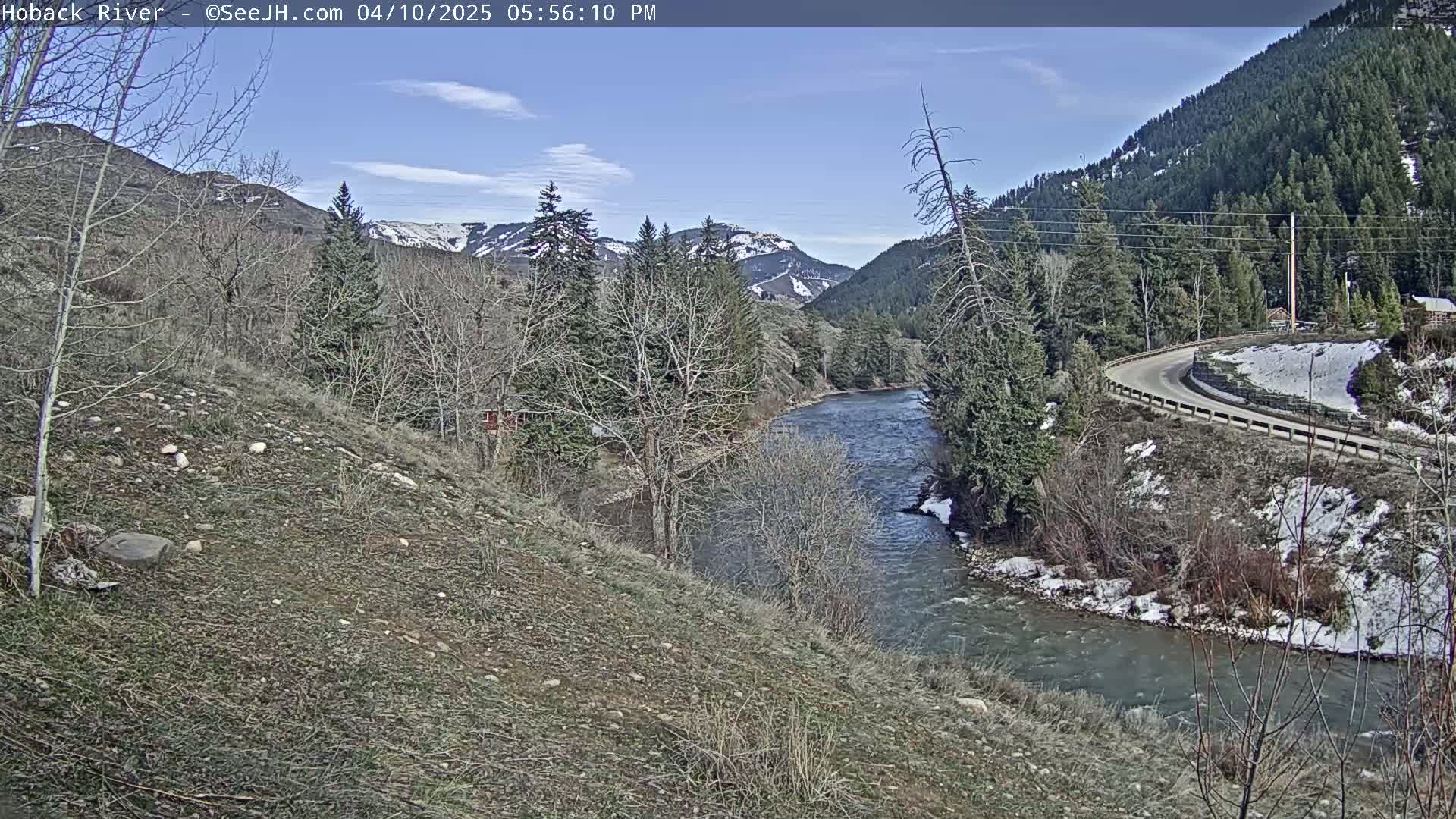 A meandering river flows through a valley on a sunny day, with snow dusting the ground and distant mountains in the background.