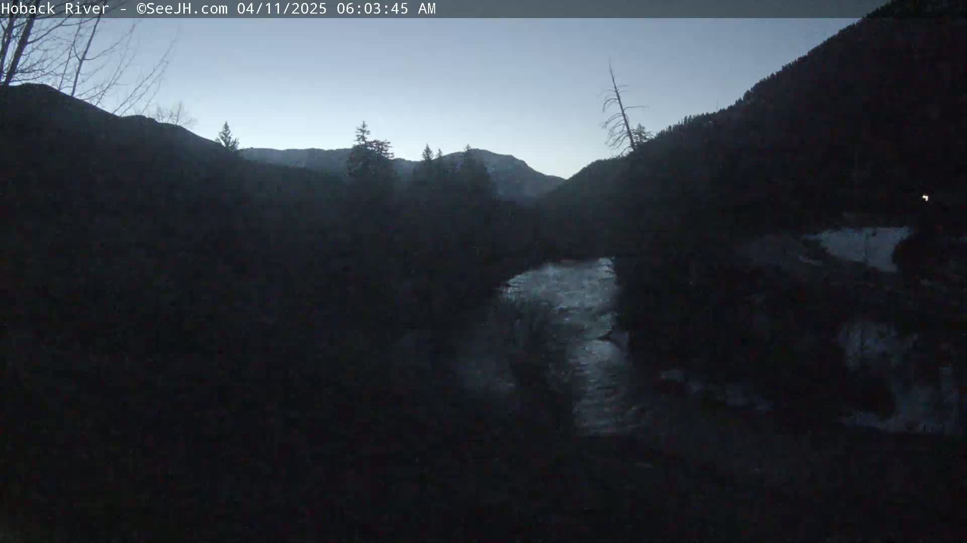 A dark, pre-dawn view of a river flowing through a mountainous valley under a twilight sky.