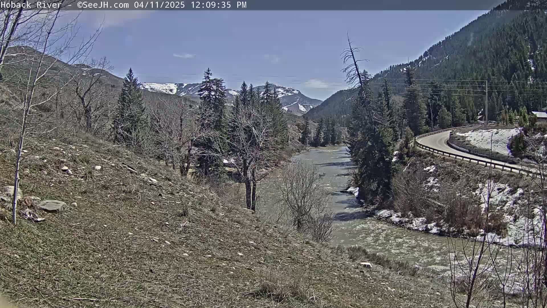 A flowing river winds through a mountain valley on a partly sunny day, with patches of snow still visible on the ground and mountainsides.