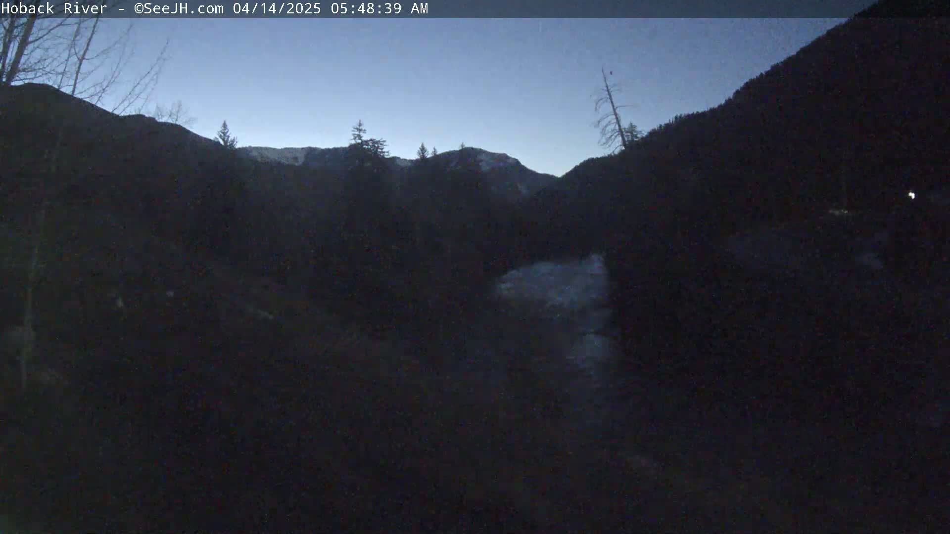 A dark, pre-dawn view of a river flowing through a mountain valley under a clear, twilight sky.