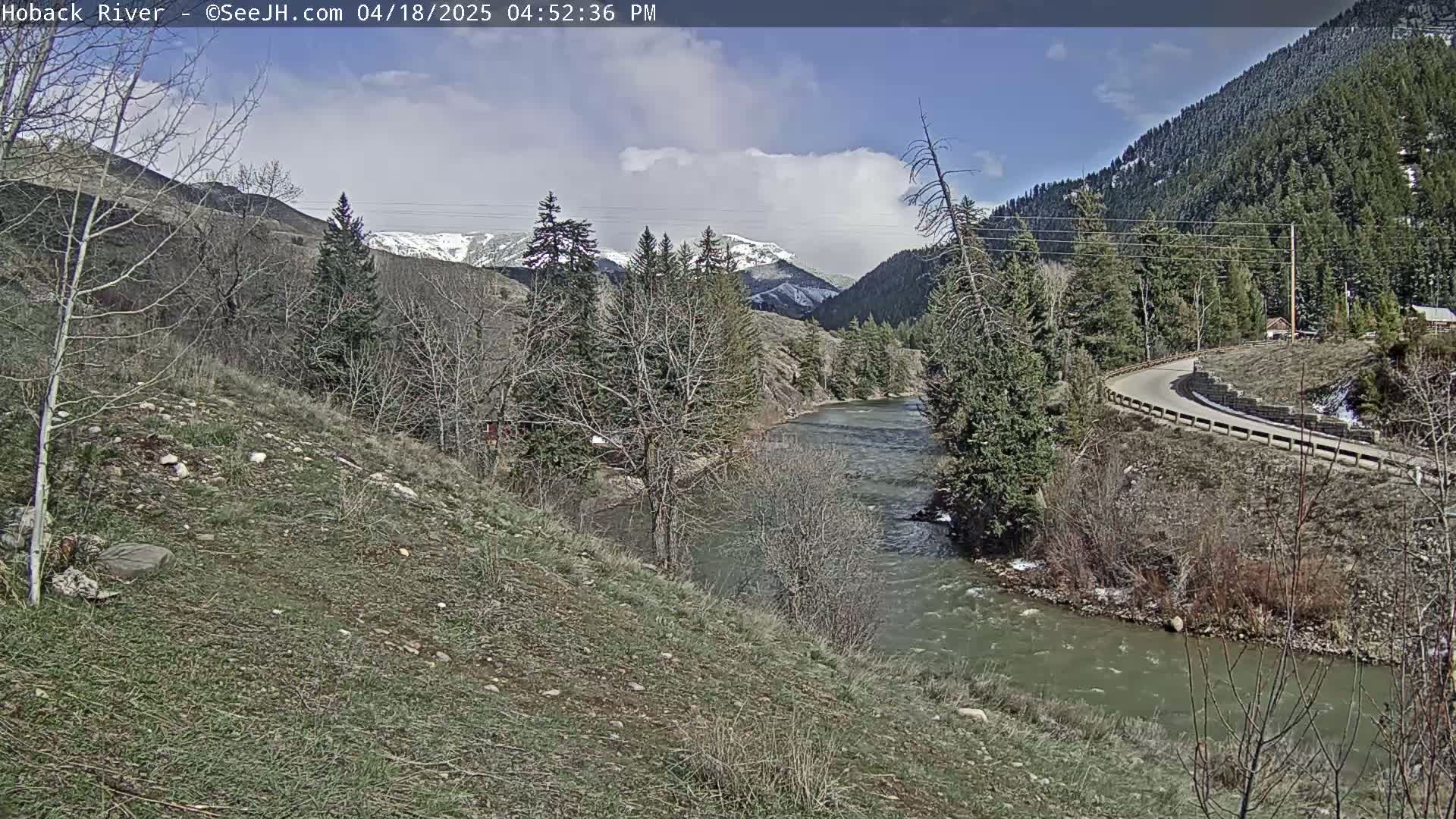 A partly cloudy day reveals a river winding through a valley, with snow-capped mountains in the distance and a paved road curving along the riverbank.