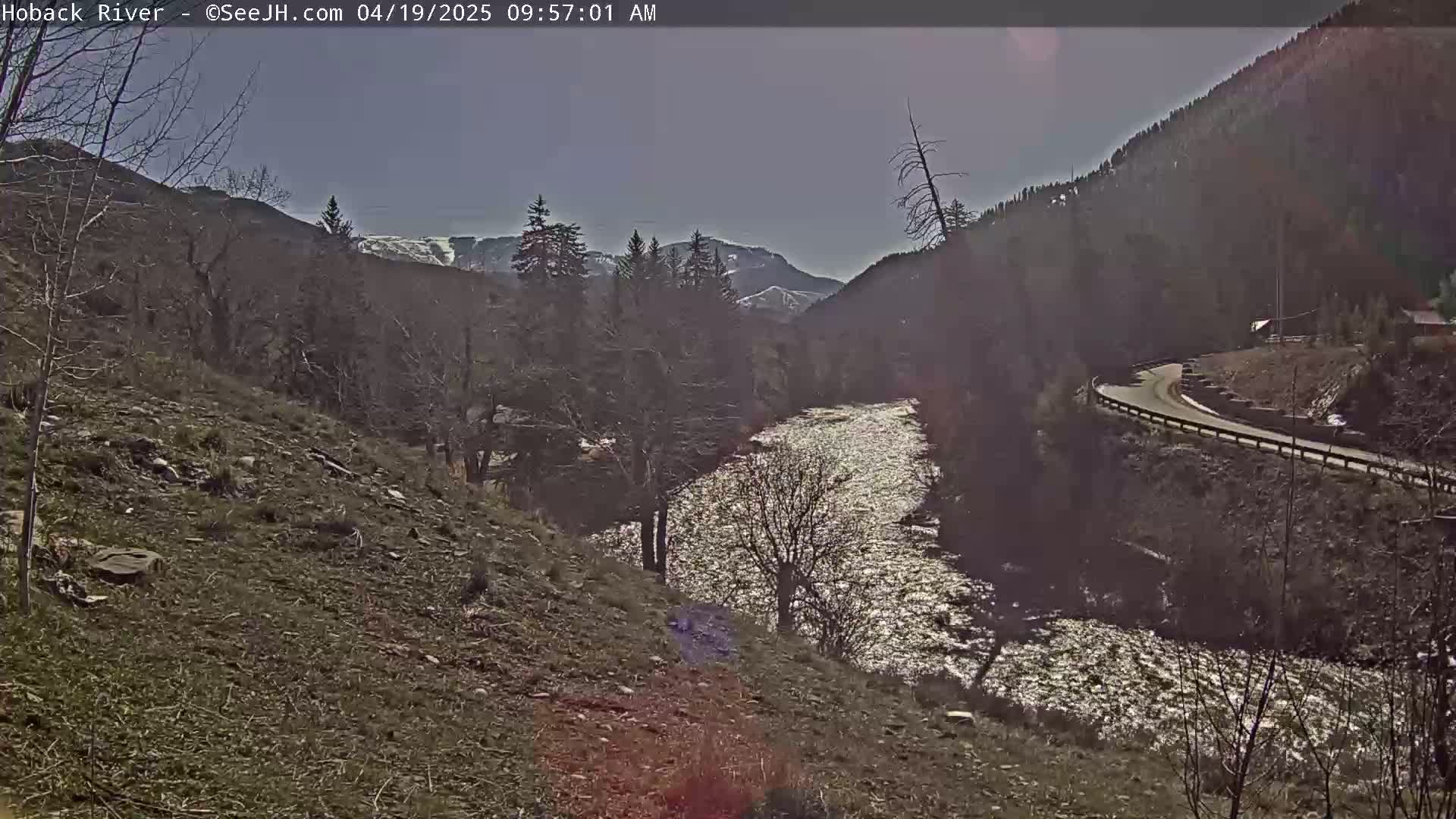 A sparkling river winds through a valley of leafless trees and rocky hillsides under a bright, clear sky.