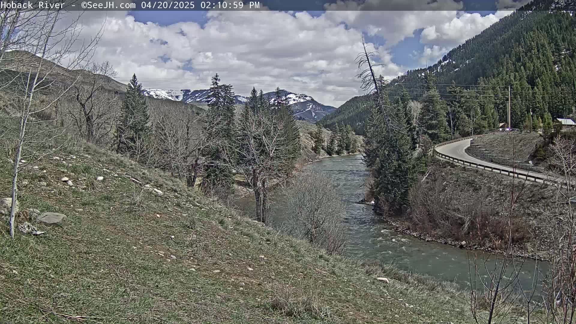 A partly cloudy day reveals a winding river flowing through a mountainous valley with a road curving along its bank, and snow-capped peaks in the distance.