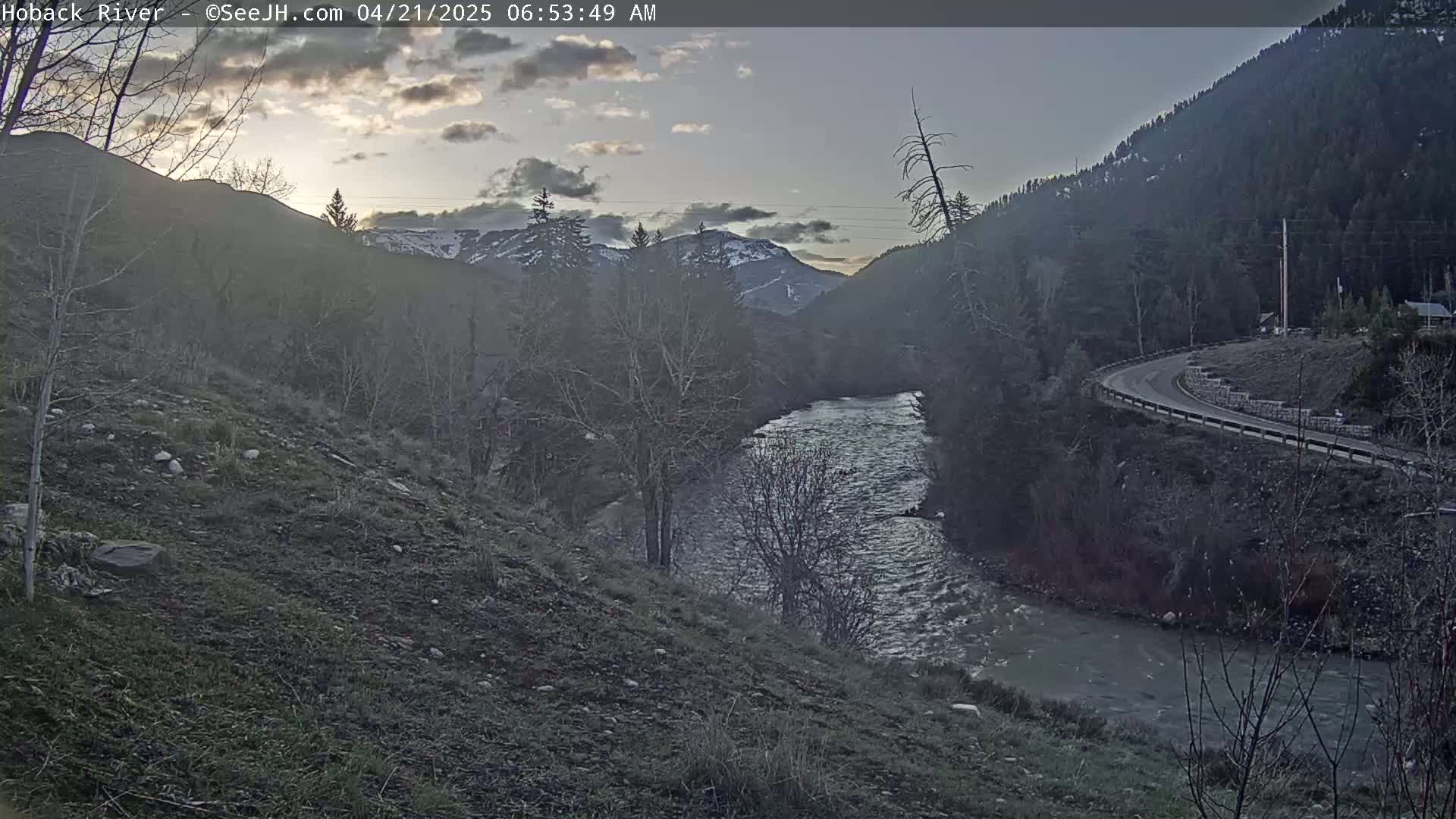 A river flows through a valley between snow-capped mountains under a partly cloudy sky at dawn.