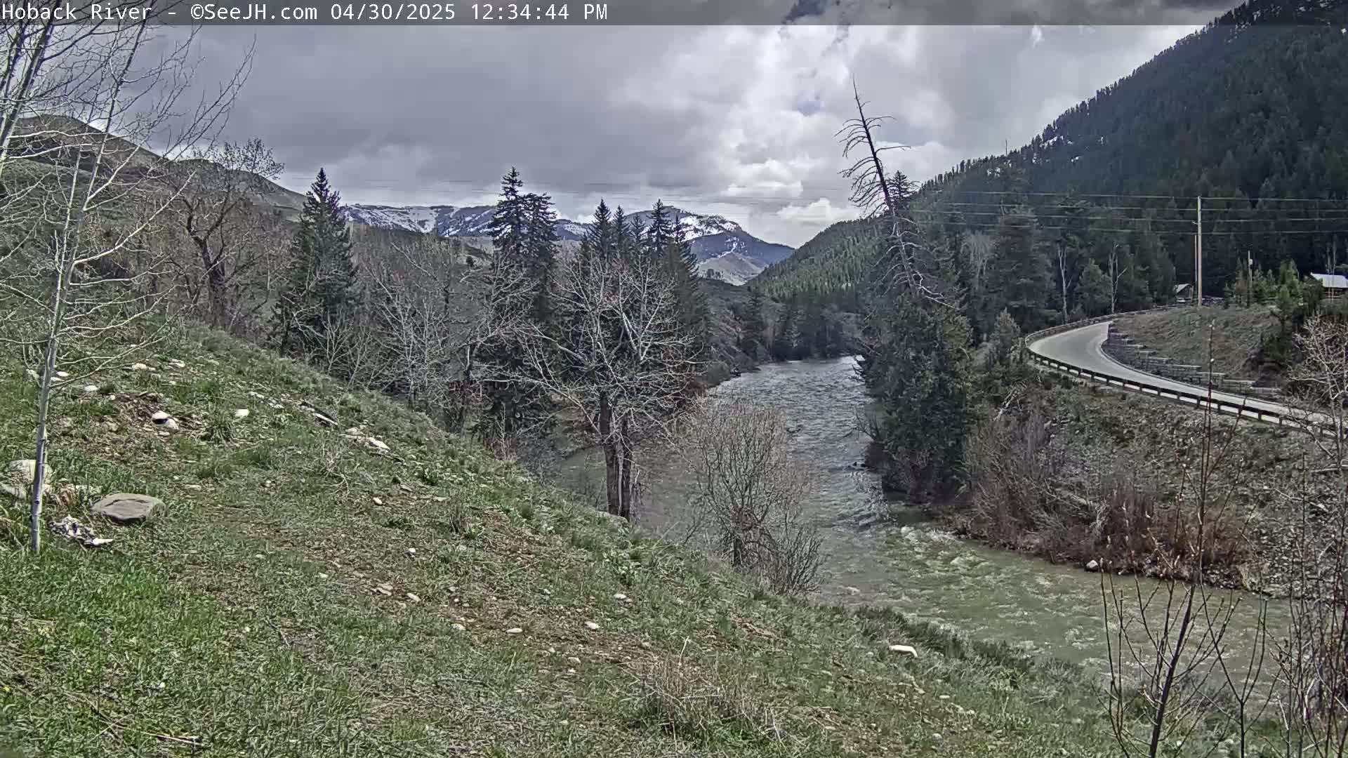 A winding river flows through a valley of leafless trees and snow-capped mountains under a cloudy sky.