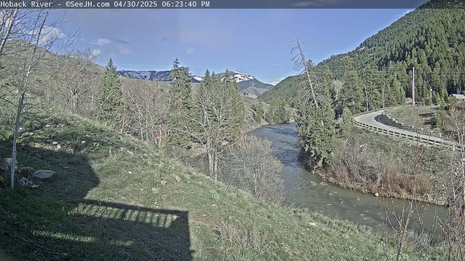 A river flows through a valley of trees and mountains under a mostly sunny sky.