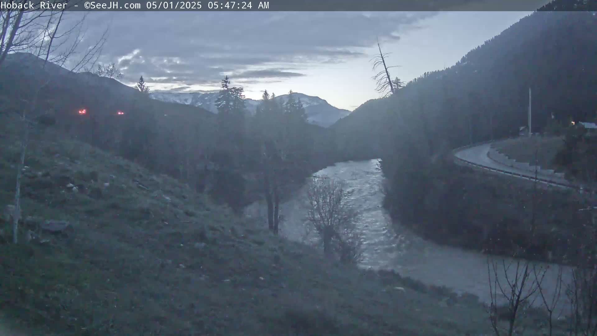 A river flows through a valley at dawn under a cloudy sky, with a road curving along the opposite bank.