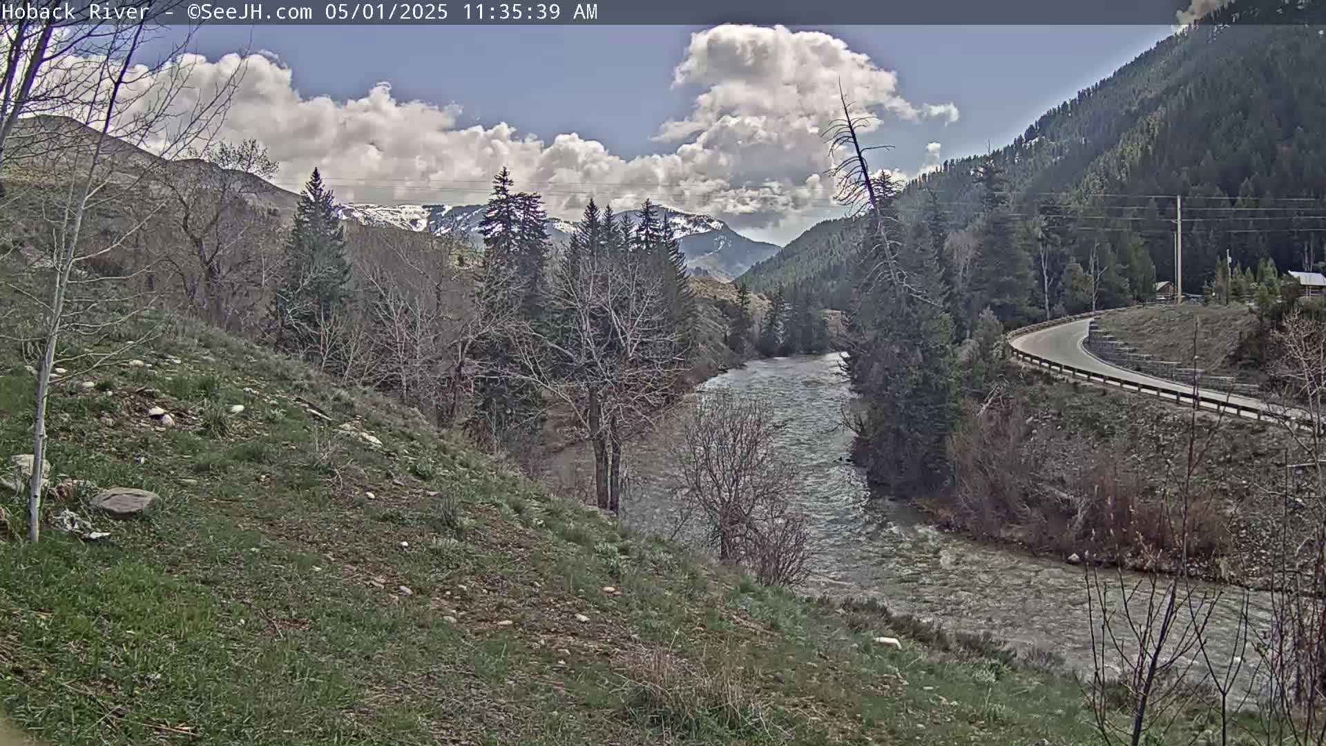 A partly cloudy day shows a river flowing through a valley, alongside a road curving through sparsely wooded hills with snow-capped mountains in the distance.