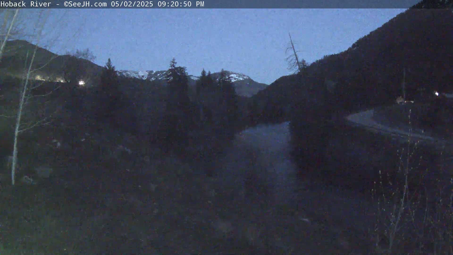 A dark, nighttime view of a river flowing through a valley, surrounded by snow-capped mountains under a twilight sky.