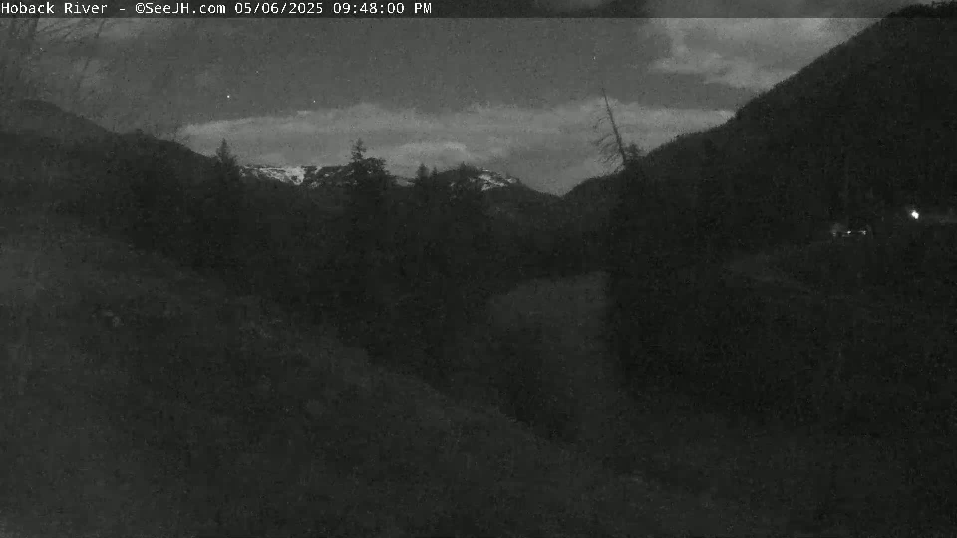 A nighttime, low-light view shows dark hills and trees under a mostly cloudy sky.
