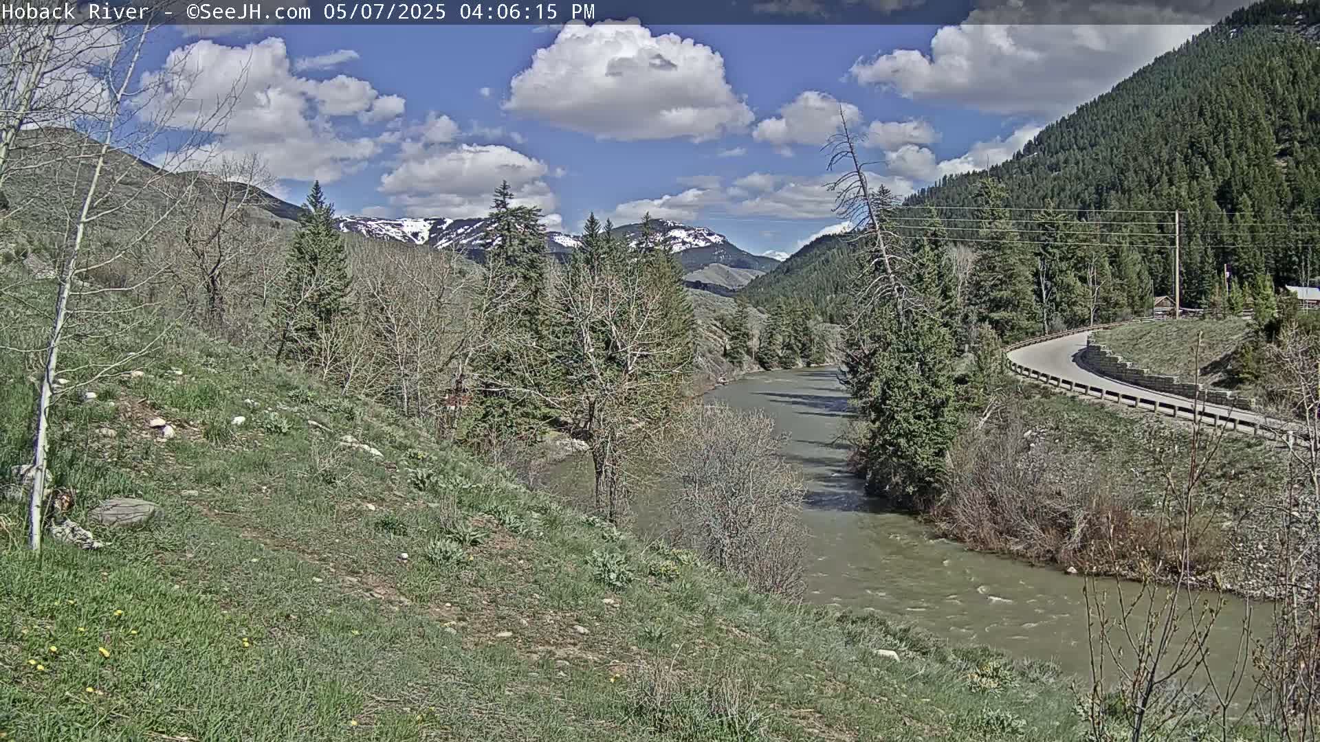 A partly cloudy day reveals a river winding through a valley of green hills and trees, with snow-capped mountains in the distance.