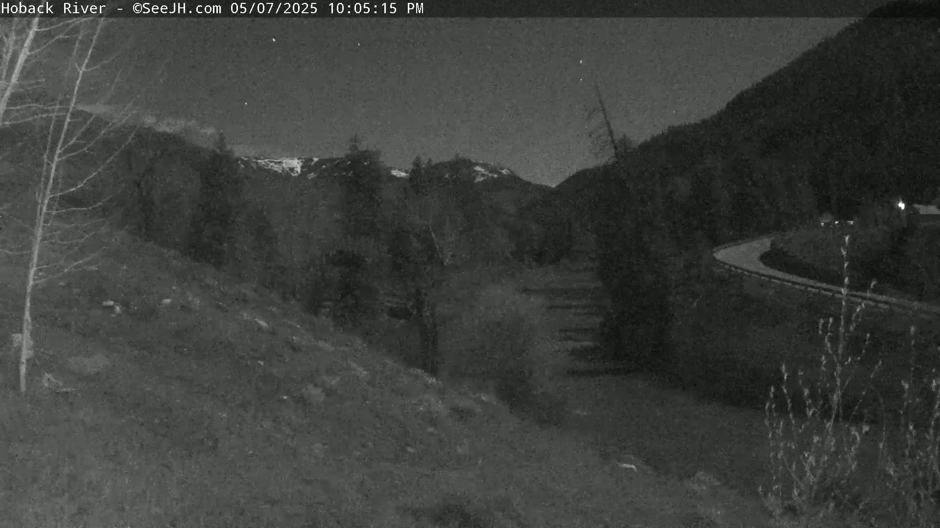 A nighttime, low-light view of a hilly landscape with sparse vegetation, a winding road in the distance, and a dark sky with a few visible stars.