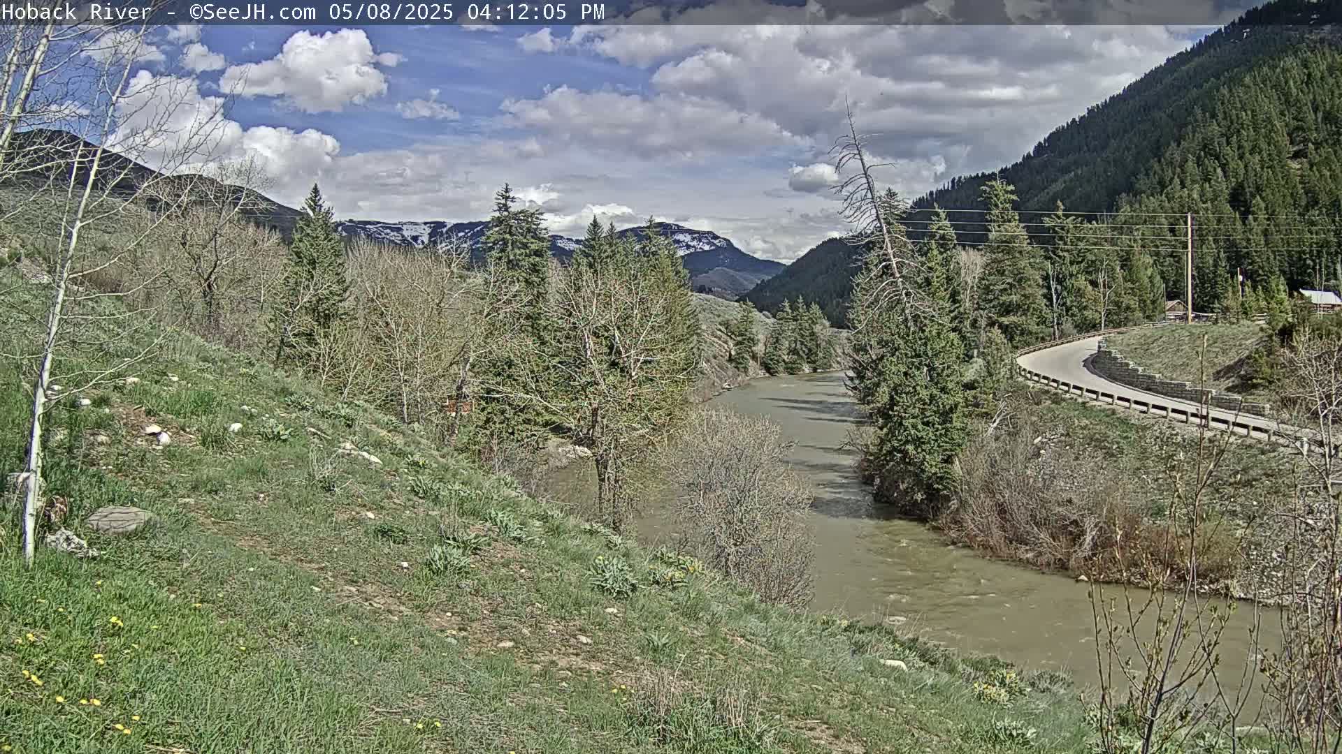A partly cloudy day reveals a winding river flowing through a valley, bordered by green hillsides, sparse trees, and a curving road.